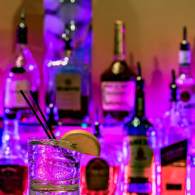 A clear drink with two straws and a lemon wedge is placed on top of a bar. In the background, an assortment of liquor bottles can be seen.
