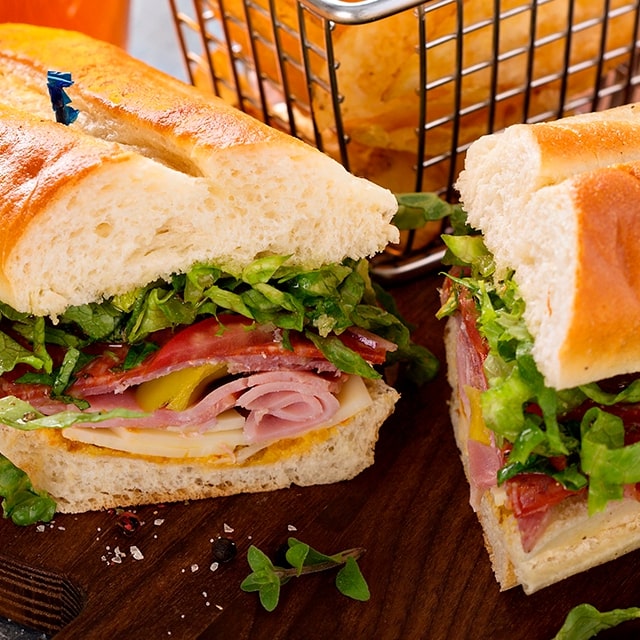 Two halves of a deli sandwhich with lettuce, tomato, meat, cheese and more are set on a table in front of a basket of chips.