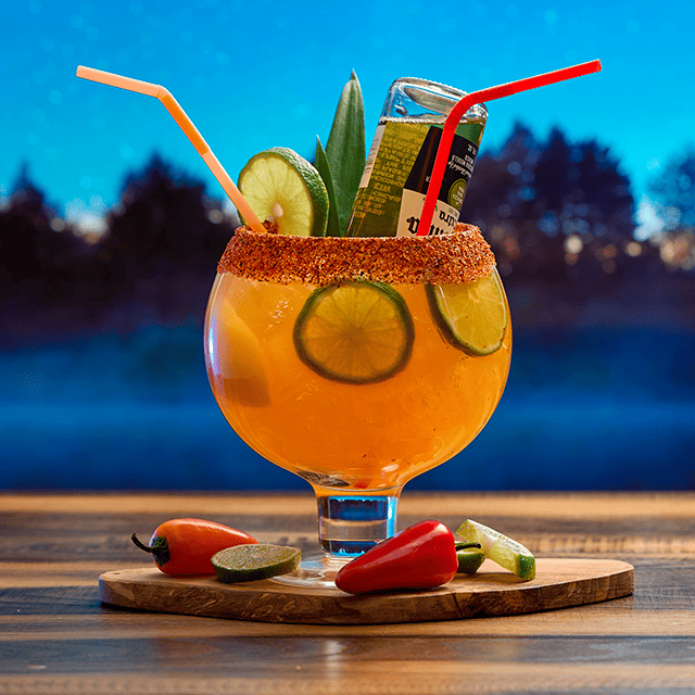 A large goblet of orange alcohol with lemon slices and chili powder on the rim is featured prominently on a wood surface. There are two straws in it, as well as an upside-down bottle.