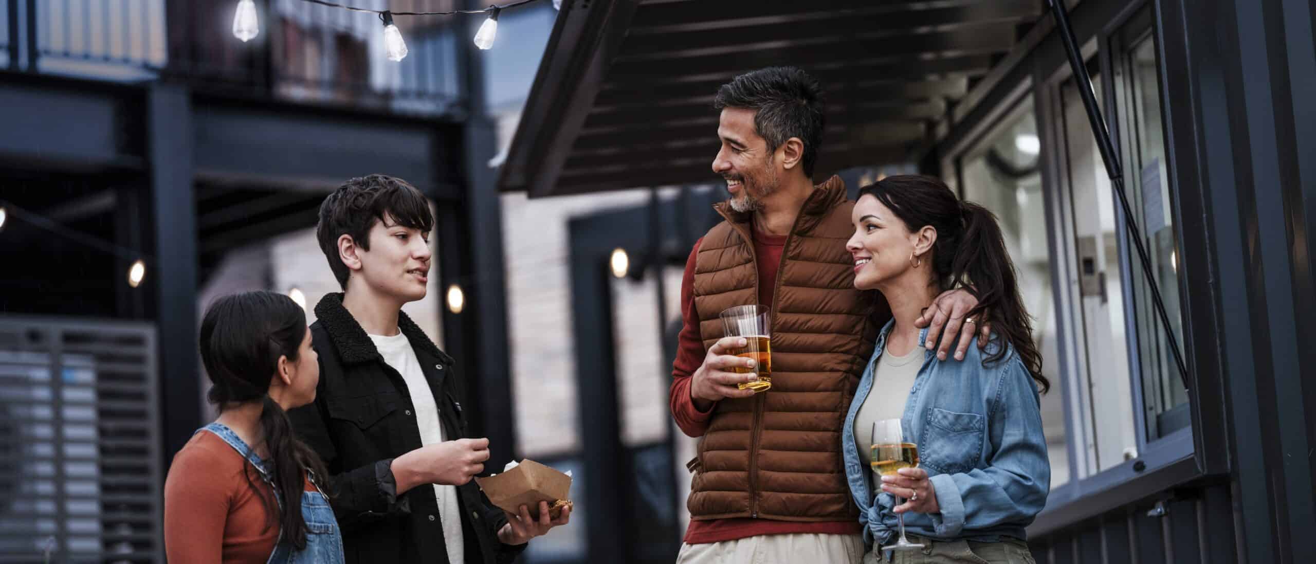 Four people stand outside, holding drinks and food and talking with each other.