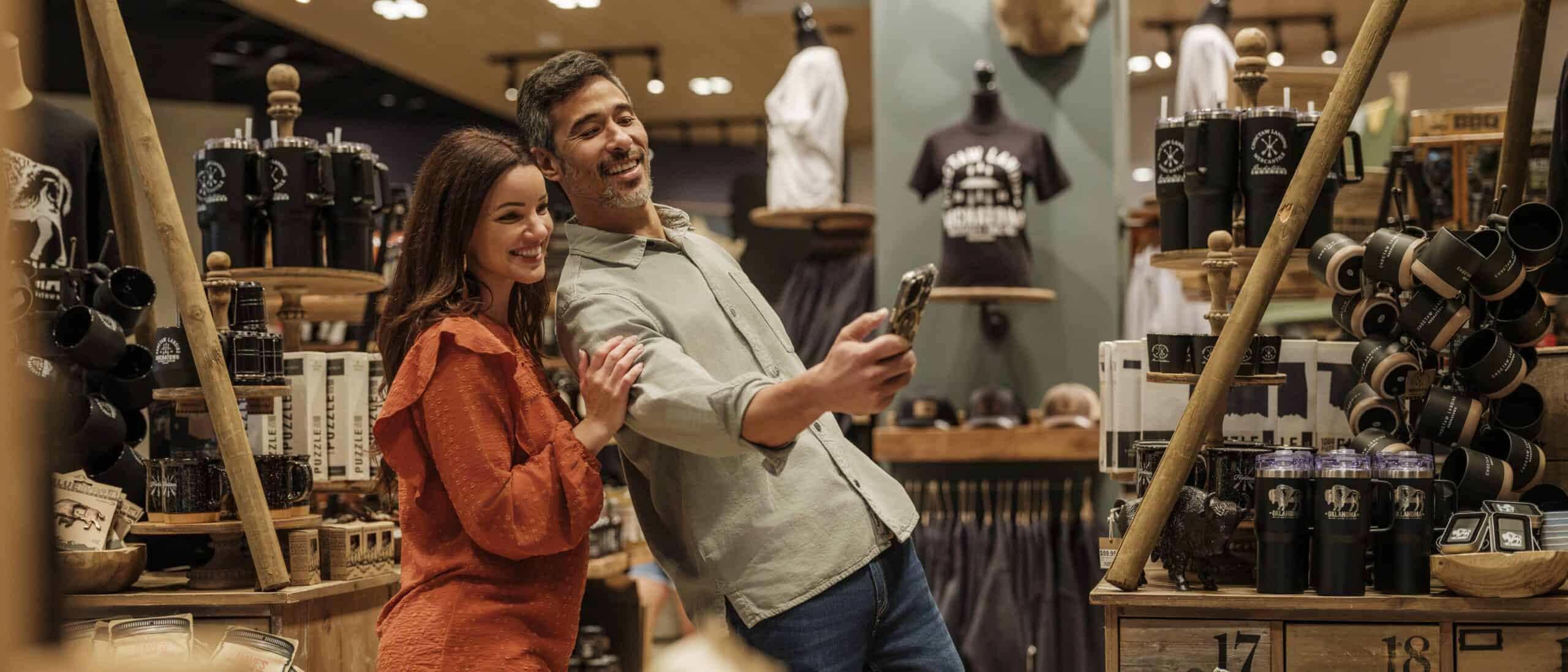 A man and woman stand in the mercantile. The man, leaning back, is showing the woman something on his phone.