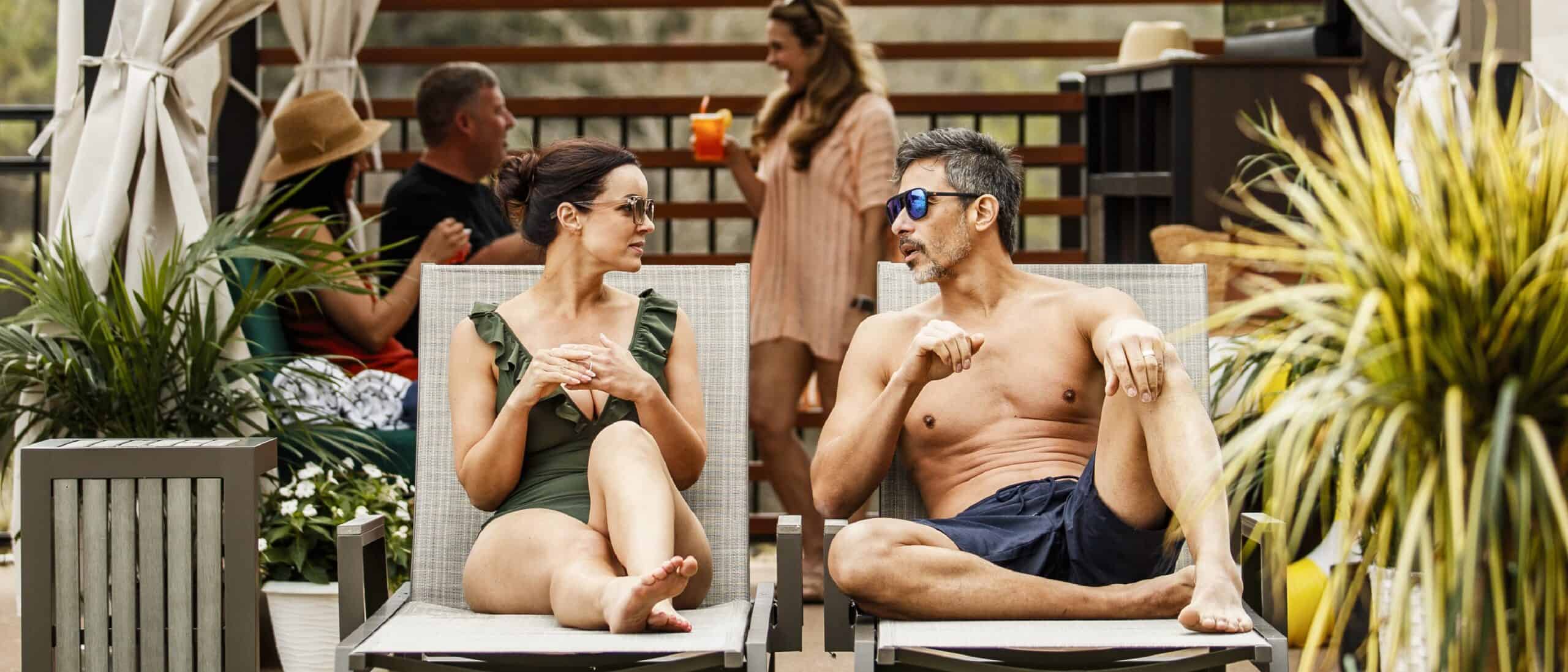 A man and woman, wearing swimsuits and sunglasses, sit in cabana chairs.