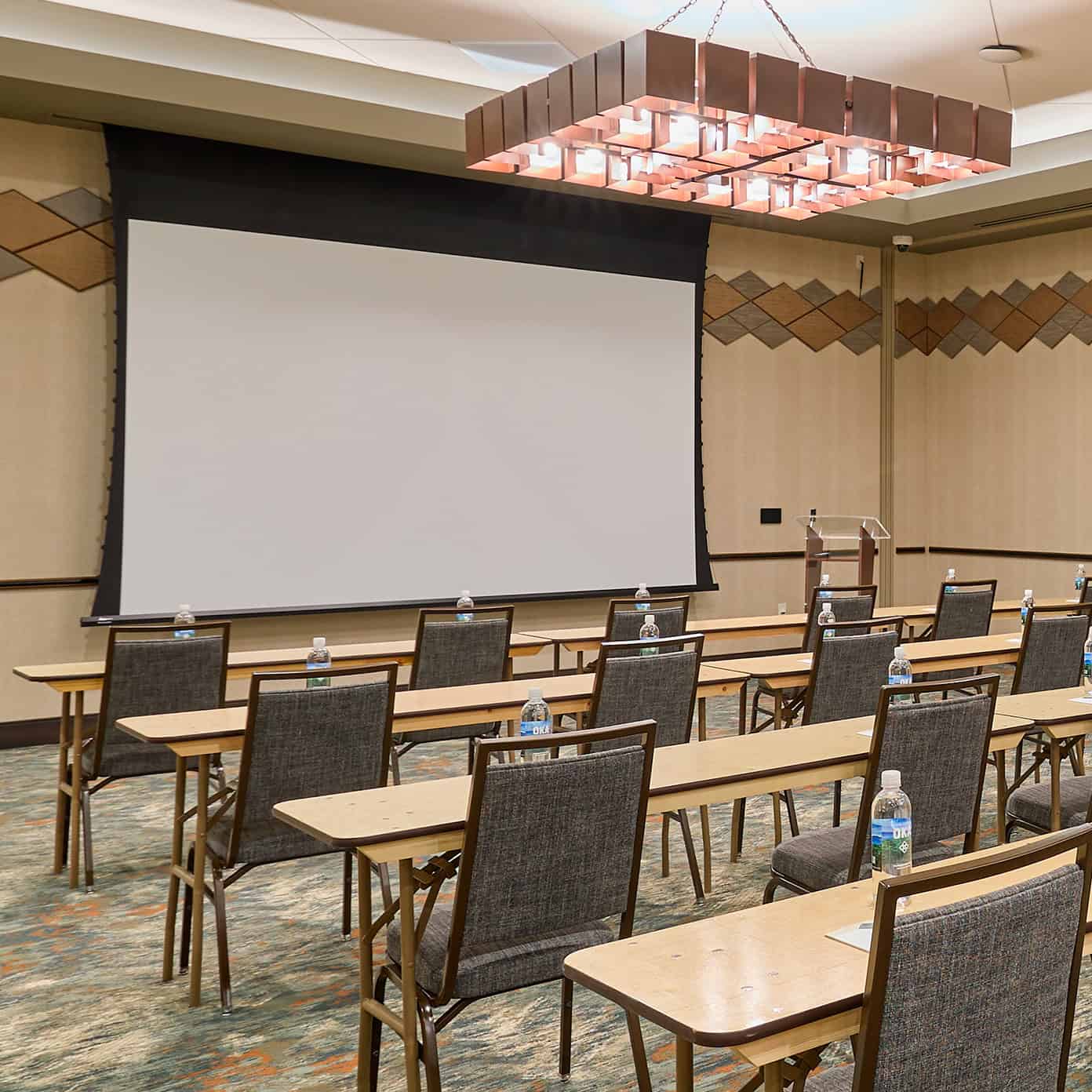 a meeting space is set up with chairs and tables in a row and a projector screen lowered in the front of the room.