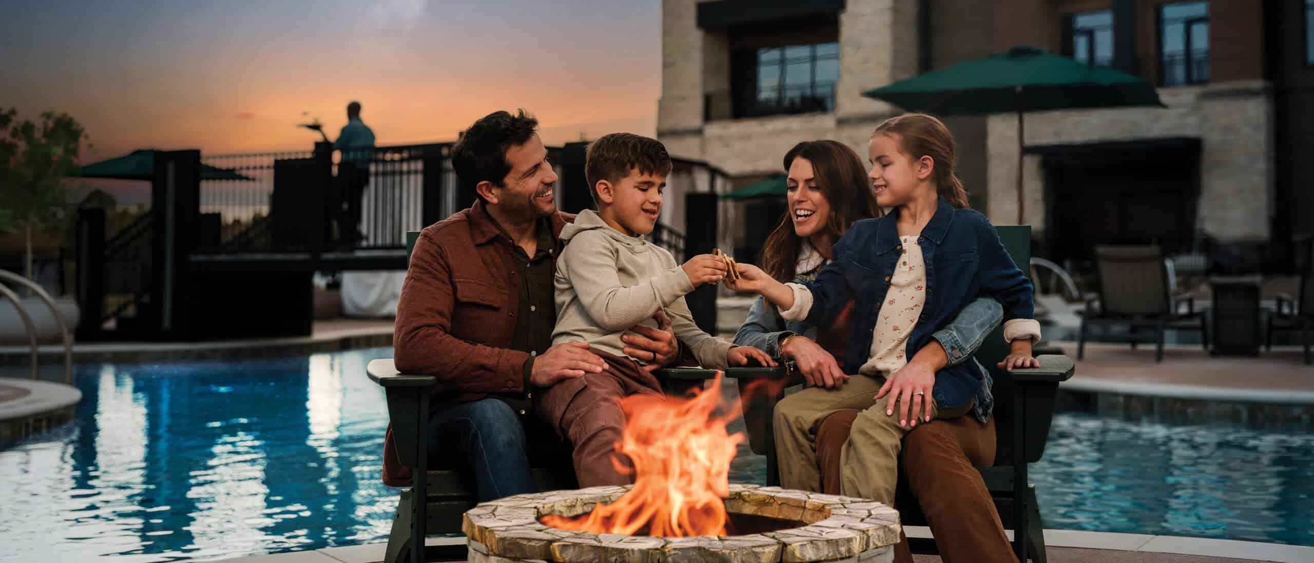 A couple and their kids sit in front of a fireplace eating s'mores at the Oka Sita pool