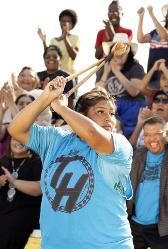 choctaw female in a blue shirt playing stickball while a crowd watches.